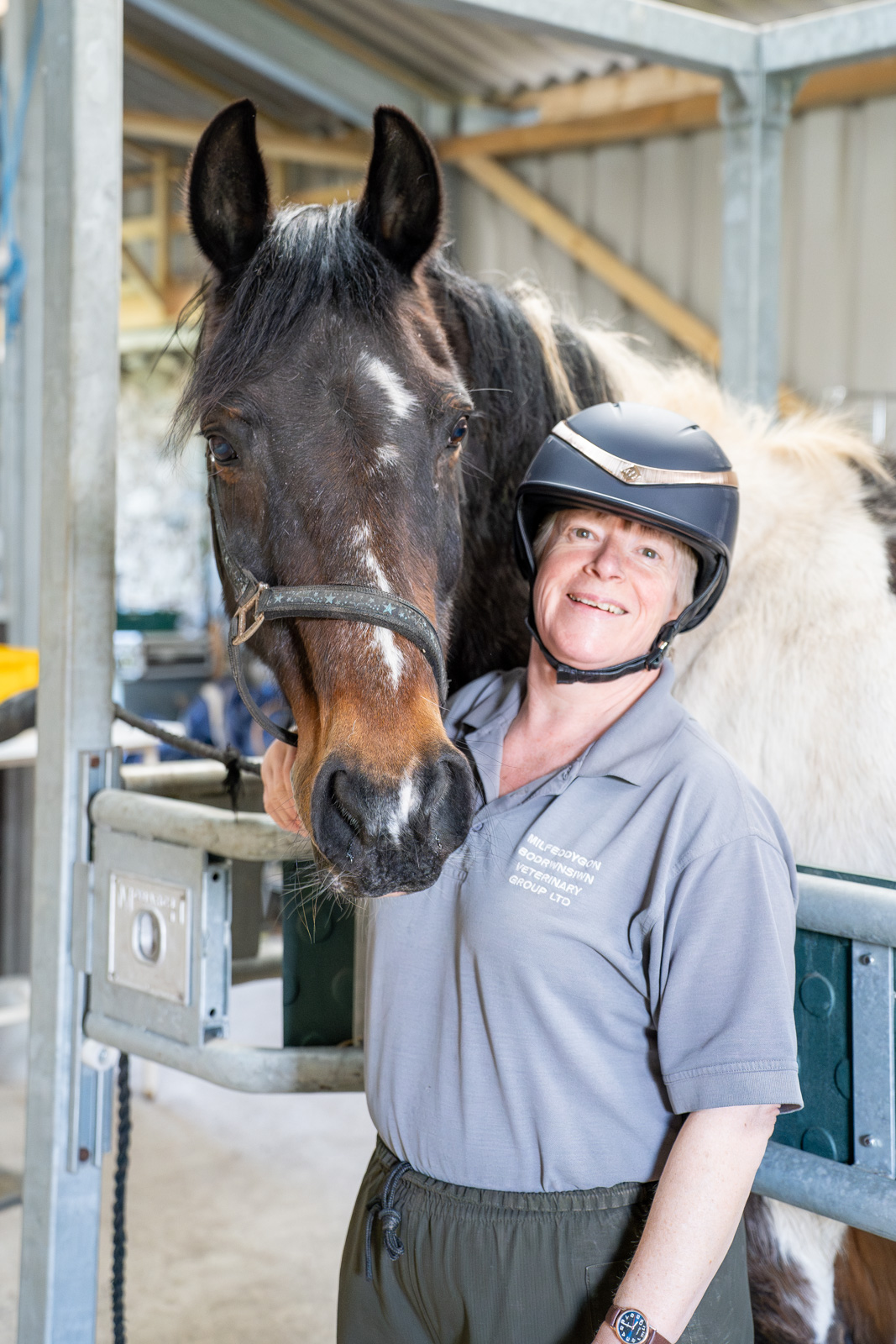 Bodrwnsiwn (Web) 162 A person wearing a helmet and gray shirt is smiling while standing next to a horse in a stable. The horse has a dark mane and bridle. The setting appears to be a well-lit barn or indoor area.