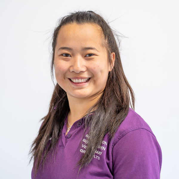 A person with long dark hair is smiling and wearing a purple collared shirt with white text. The background is plain and light-colored.